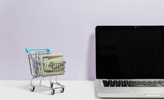 macbook pro on white table beside a miniature shopping cart with money